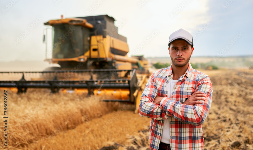 Happy farmer proudly standing in a field with arms crossed on chest. Combine harvester driver going to crop rich wheat harvest. Agronomist wearing flannel shirt, looking at camera on a farmland.