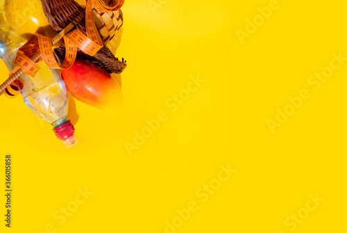A basket of fresh fruit: a red Apple, a yellow pear and a mango lie surrounded by a measuring tape on a yellow background next to a sports water bottle: a healthy food concept, place for text, top 