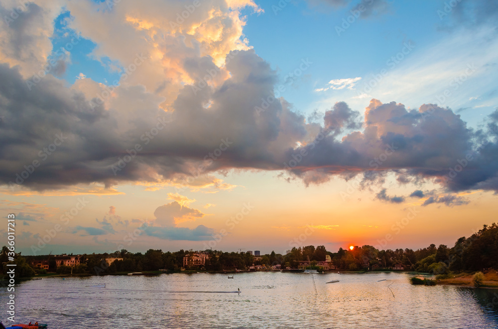 Evening sky in blue and pink tones, with curly clouds, illuminated by the setting sun over the surface of the lake with a distant wooded shore and urban buildings.