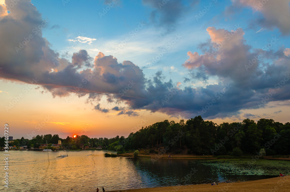 Evening sky with the setting sun and a strip of yellow sand on the shore of the city lake with vacationers. Evening sky with the setting sun and a strip of yellow sand on the shore of the city lake with vacationers.