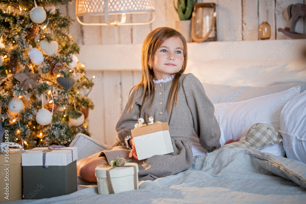 portrait of a child girl smiling, sitting on a bed with a gift in her hands, in the background in blurred focus a Christmas tree glows with garlands, bokeh portrait of a child girl smiling, sitting on a bed with a gift in her hands, in the background in blurred focus a Christmas tree glows with garlands, bokeh