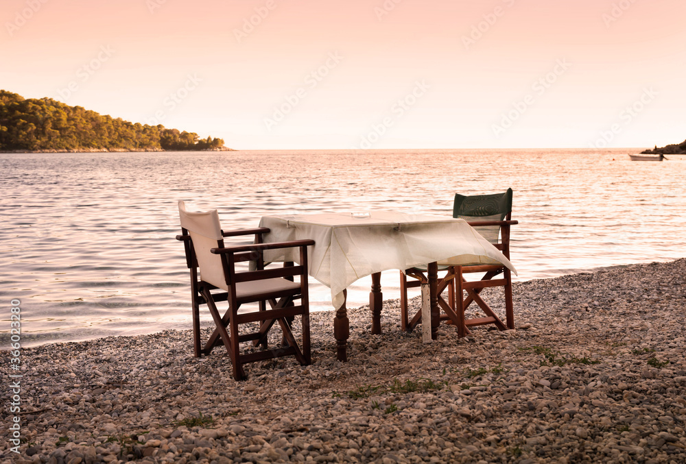 A table and two chairs stand on the beach near the Mediterranean sea in warm evening sun, waiting for a romantic dinner.