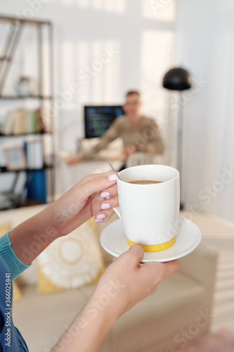 Hands of wife bringing cup of coffee to her husband who is working on computer at home due to pandemic