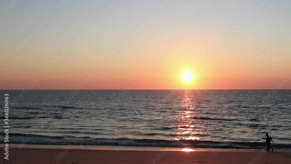 Slow Motion panoramic view of Silhouette unrecognized people walking golden sandy beach at evening twilight sunset with reflection on sea water surface & calm ocean waves in cloudsless red pink sky Slow Motion panoramic view of Silhouette unrecognized people walking golden sandy beach at evening twilight sunset with reflection on sea water surface & calm ocean waves in cloudsless red pink sky