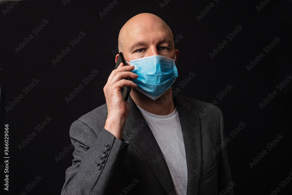 Portrait of a bald businessman in white t-shirt and grey suit on dark background, talking on black smart phone and wearing blue protective face mark during COVID 19 flue outbreak. Portrait of a bald businessman in white t-shirt and grey suit on dark background, talking on black smart phone and wearing blue protective face mark during COVID 19 flue outbreak.