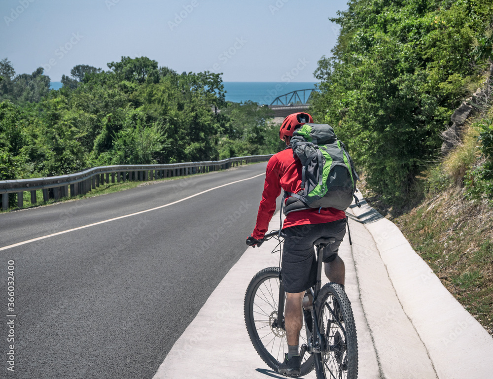 Active man with backpack riding bicycle on roadside of asphalt road at sunny summer day