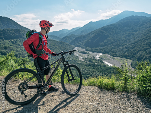 Man with hydration pack and water bladder standing with cross country bike and looking at mountains and river