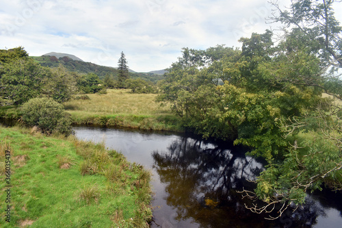 Rustic scene in the Welsh countryside near to the Snowdonia National Park. A river meanders gently through the fields on a summers day. Trees line the bank, reflecting on the still waters.