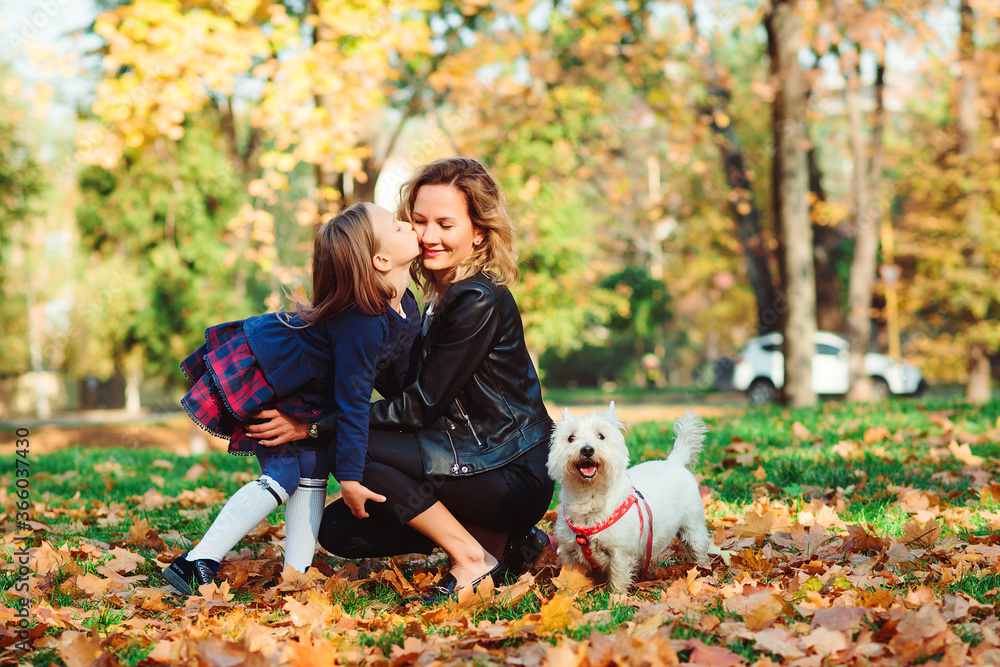 Fototapeta premium Fashionable mother and schoolgirl walking with their dog in park. Autumn time, weekend and holidays.