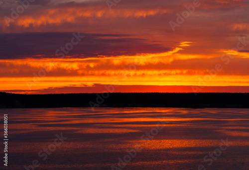 A fiery sunset is reflected in the calm river water  overhanging clouds in the bright orange sky  light waves on the water surface.