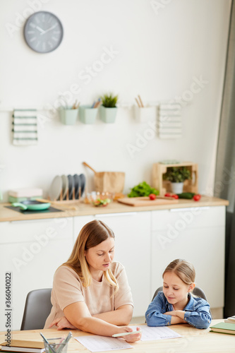 Vertical warm-toned portrait of caring mother helping daughter doing homework and studying at home in cozy interior, copy space