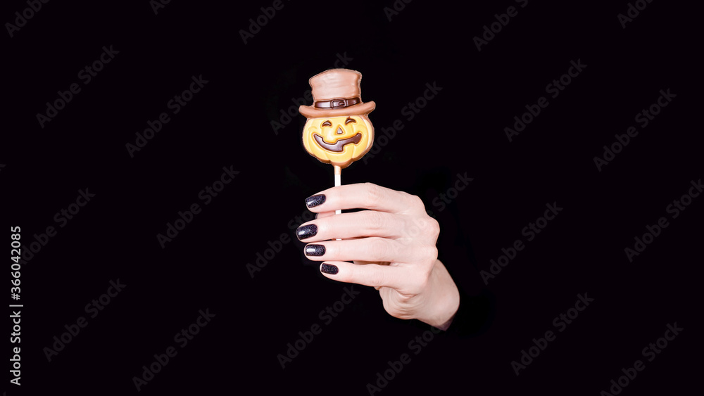 Female hand holding chocolate candy little pumpkin on the stick, black panorama background. Copy space
