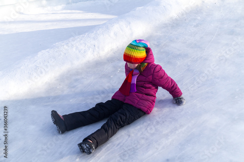 Child rolls down an ice slide. Bright clothes rainbow hat.