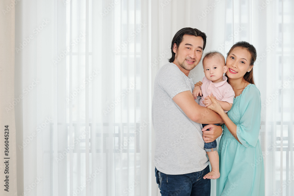 Happy young parents holding their serious little son and looking at camera Happy young parents holding their serious little son and looking at camera
