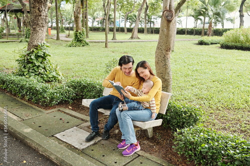 Happy young parents sitting on bench in park and reading interesting book to their little son Happy young parents sitting on bench in park and reading interesting book to their little son