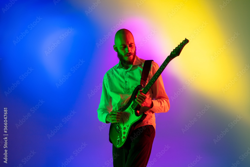 Tensioned. Young caucasian inspired and expressive musician, guitarist performing on multicolored background in neon. Concept of music, hobby, festival, art. Joyful artist, colorful, bright portrait.
