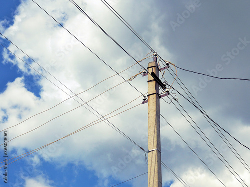 Electric concrete pillar against a blue sky. Illustration on the theme of electricity supply and rising prices. The wires cross against a background of fluffy white clouds. Rural electrification