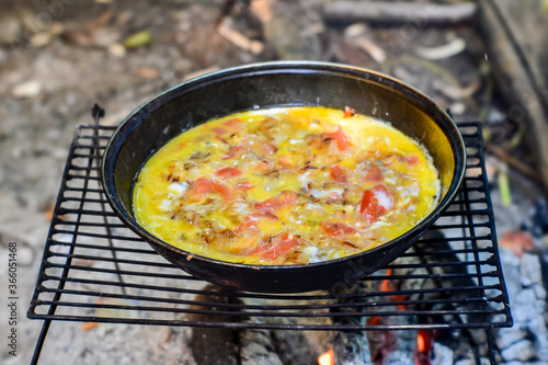 Fried eggs with vegetables are cooked in a pan on a grill on coals in the open air.