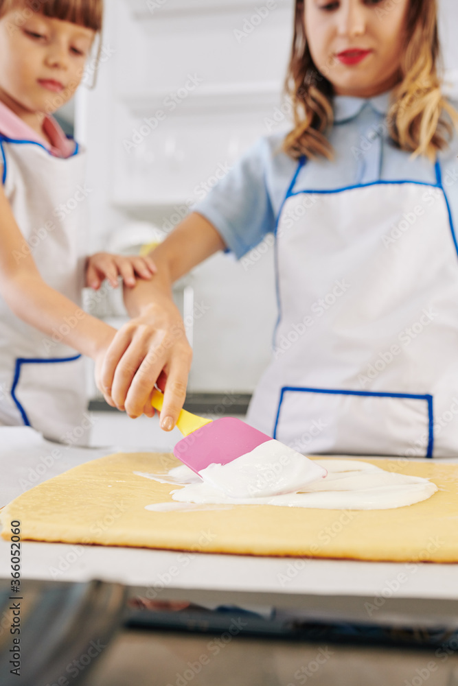 Mother helping daughter to apply whipped cream on cake when they are making swiss roll at home Mother helping daughter to apply whipped cream on cake when they are making swiss roll at home