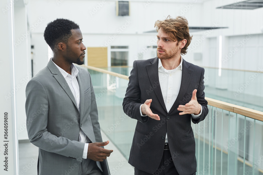 Waist up portrait of two successful businessmen wearing suits chatting while walking toward camera in modern office building interior Waist up portrait of two successful businessmen wearing suits chatting while walking toward camera in modern office building interior