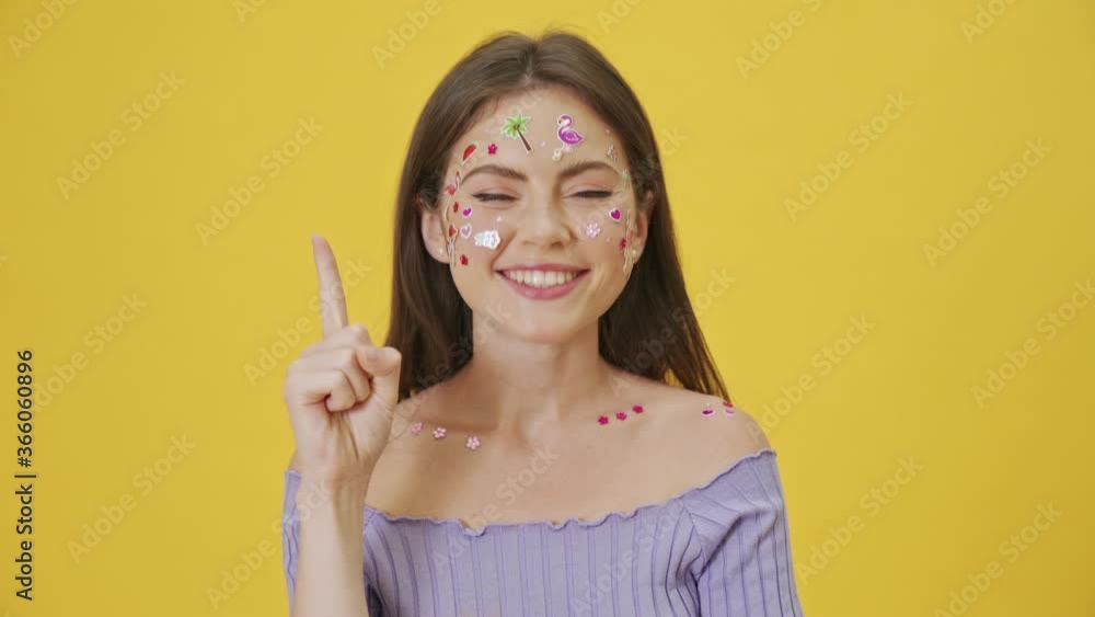 A serious young girl with fashion makeup and stickers on the face is thinking about something then come-up with an idea standing isolated over a yellow background A serious young girl with fashion makeup and stickers on the face is thinking about something then come-up with an idea standing isolated over a yellow background