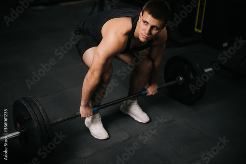 Young muscular with perfect beautiful body wearing sportswear lifting heavy barbell from floor during sport workout training in modern dark gym. Concept of healthy lifestyle.