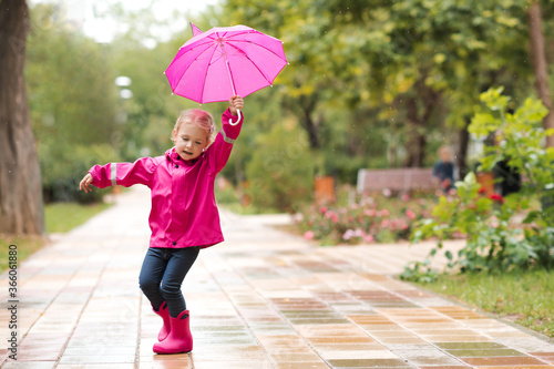 Kid girl 2-3 year old walking on puddle in park holding pink umbrella wearing waterproof boots outdoors. Childhood. Autumn season.