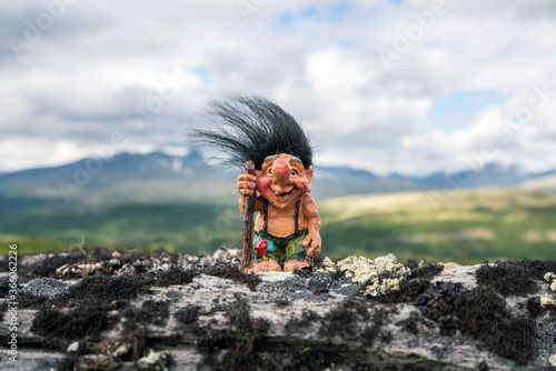 Funny norwegian troll figure with big nose and walking stick outdoors in the mountains. Hair standing up because of windy conditions.