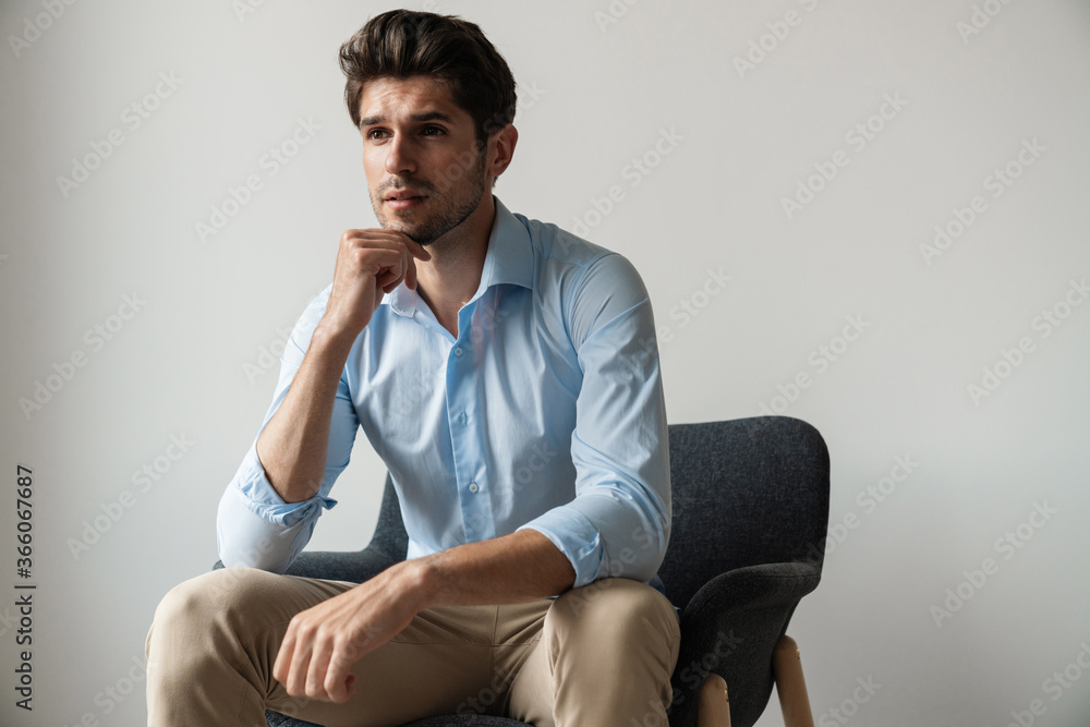 Image of serious brunette man thinking and sitting on chair in room Image of serious brunette man thinking and sitting on chair in room