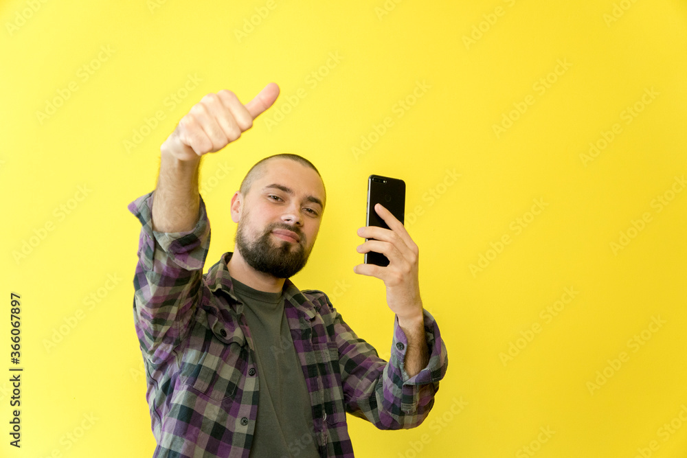 Young smiling caucasian man with a beard and a bald head holding a phone in his hands on a yellow background. Approval, thumbs up, like. The guy recommends. Copy space Young smiling caucasian man with a beard and a bald head holding a phone in his hands on a yellow background. Approval, thumbs up, like. The guy recommends. Copy space