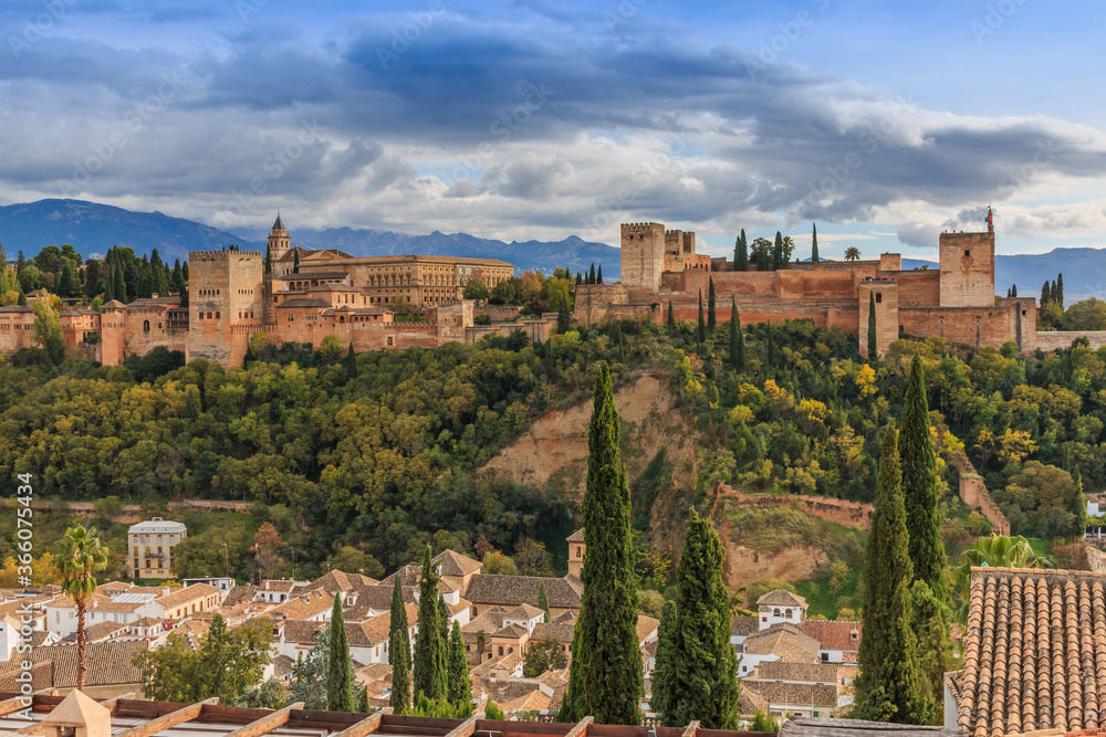 Spanish city of Granada in Andalusia with clouds and mountains in the background. Historic Alhambra fortress with trees and buildings on a hill. Red tile roofs of the buildings to the autumn mood