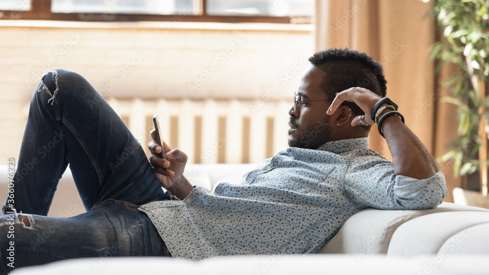 Young african American man in glasses lying resting on comfortable sofa in living room using smartphone gadget, biracial male relax on couch at home browse internet or texting on cellphone at home Young african American man in glasses lying resting on comfortable sofa in living room using smartphone gadget, biracial male relax on couch at home browse internet or texting on cellphone at home