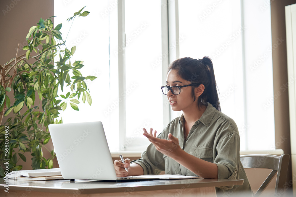 Indian woman online tutor remote teacher wearing glasses speaking to webcam  chat explaining online class zoom video call school lesson looking at  laptop virtual conference meeting work at home office. foto de