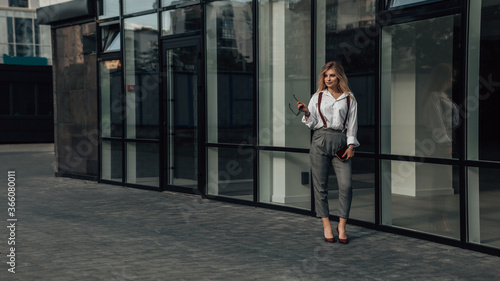 Full length portrait of young female CEO, manager, director, glass skyscraper. Business concept. Feminine gait. Gorgeous agent or entrepreneur holding sunglasses and clutch bag. Copy space.