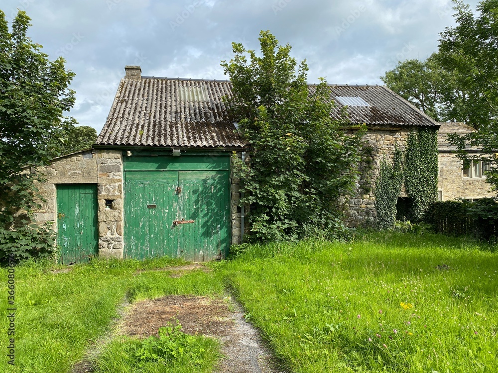 Old empty farm building, with long grass, trees, and green painted doors in, Hetton, Skipton, UK