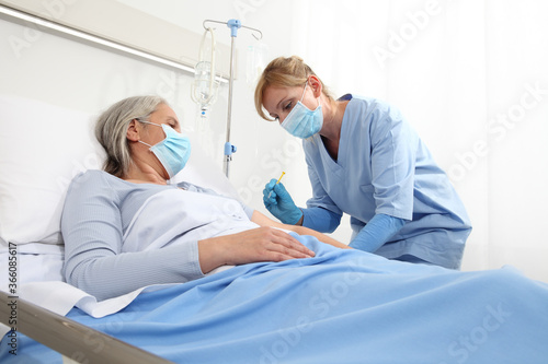 nurse with the syringe injects the vaccine to the elderly woman patient lying in the hospital room bed, wearing protective gloves and medical surgical mask, coronavirus covid 19 protection concept