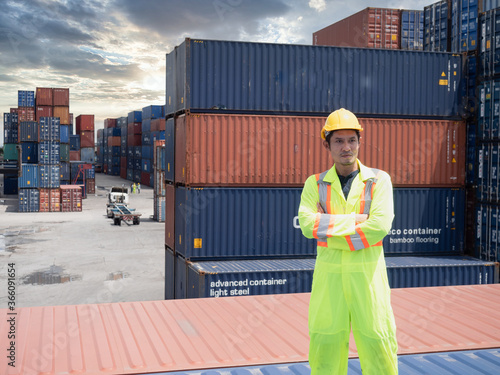 Portrait of smart engineer man wearing a safety helmet and safety vest standing and Looking at the stock container management in container yard background..