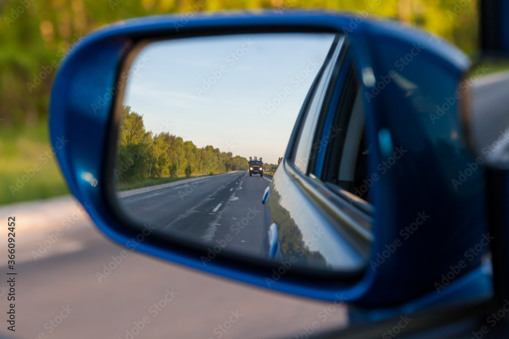 Rear view in the side mirror of a blue sedan with the reflection of a gray truck on an asphalt road on a summer day with green trees on the sides of the highway.