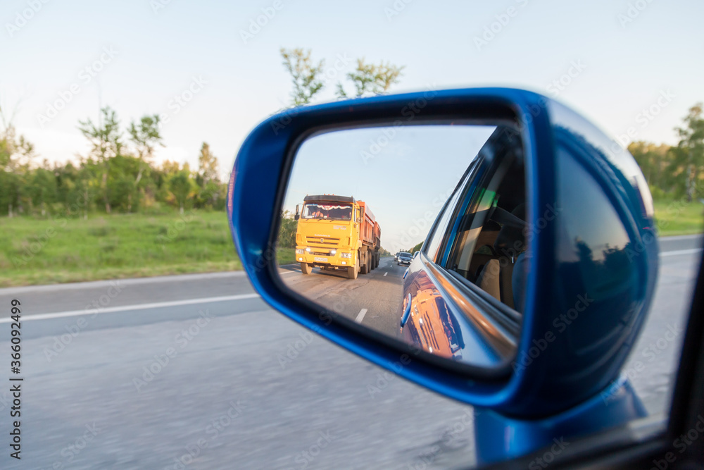 Rear view in the side mirror of a blue sedan with the reflection of an overtaking yellow KAMAZ truck on an asphalt road on a summer day with green trees on the sides of the highway. Traffic Laws.
