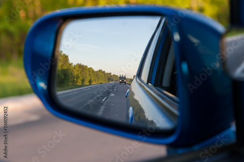 Rear view in the side mirror of a blue sedan with the reflection of a gray truck on an asphalt road on a summer day with green trees on the sides of the highway.