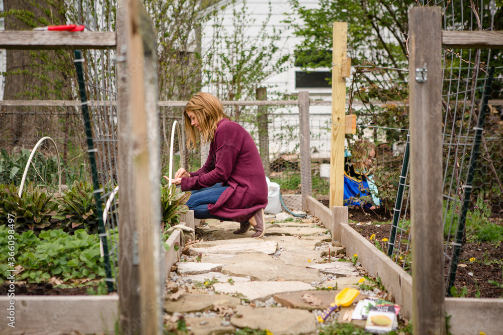 A woman plants seeds in her garden while child plays in background A woman plants seeds in her garden while child plays in background