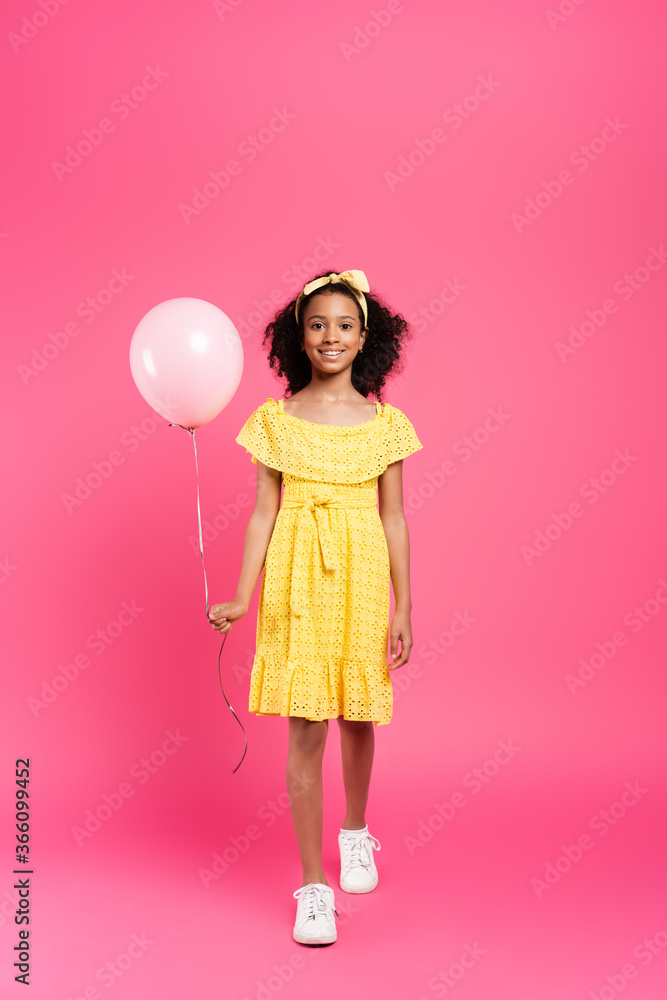 full length view of smiling curly african american child in yellow outfit with balloon on pink background