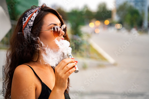 Amazing, attractive boho brunette enjoying smoking e cigarette on city street   photo