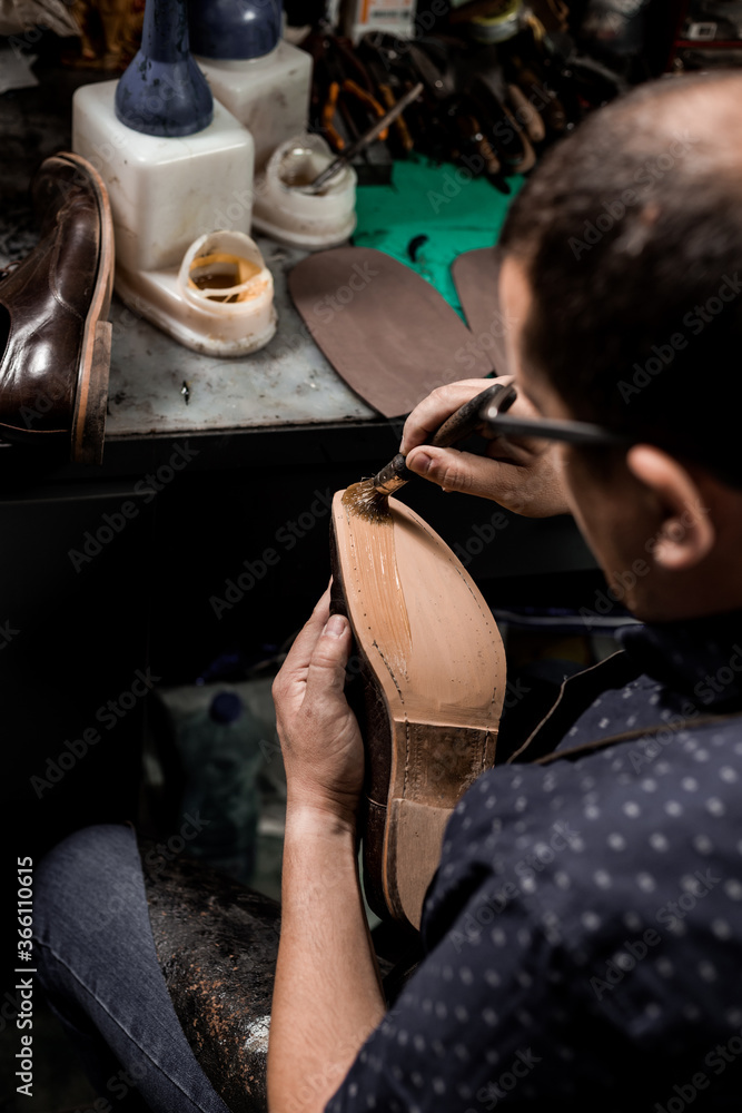 Obraz premium Male cobbler sitting at his shop and repairing man's shoe, brushing glue over sole.