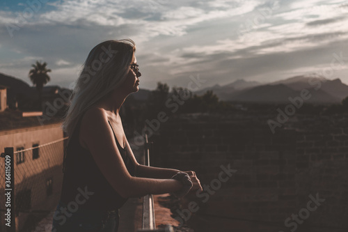 Young blonde girl watching the sunset in an old Spanish village in the summer of 2020 The background has a palm tree, mountains and white clouds. She is wearing a black tank top and black glasses.