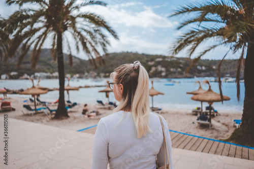 A young blonde girl walking in the port of Soller in Palma de Mallorca Spain. Summer of 2020 Her blonde and light brown hair is half up with a ponytail. Palm trees, sun chairs and white sand, mountain © Nicolas