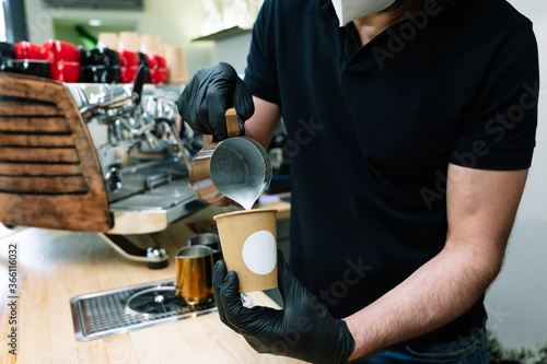 Waiter preparing coffee.Pouring milk with black gloves.He wears a mask on his face.