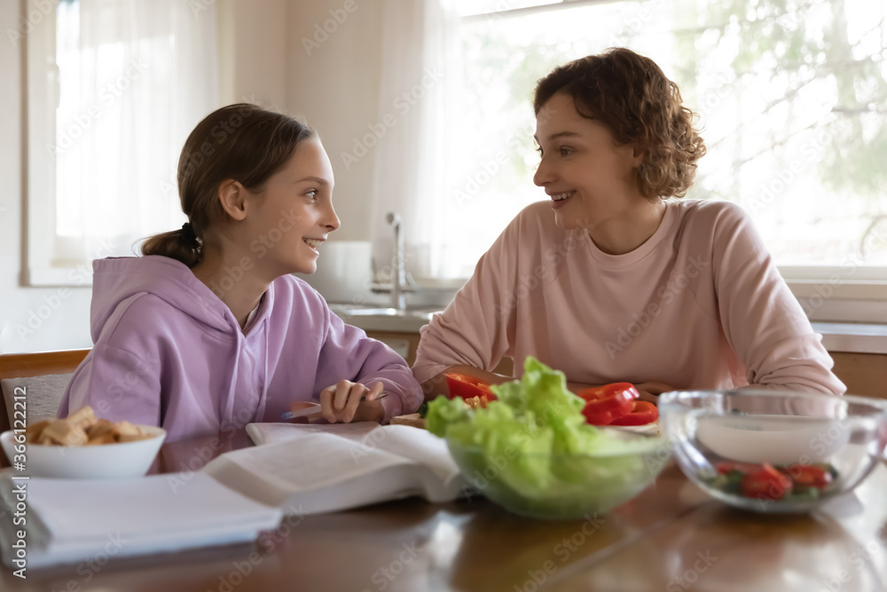Happy mother and teenager daughter chatting, sitting at table in modern kitchen, smiling teen schoolgirl studying, working on school assignments, homework, mum cooking salad, preparing dinner