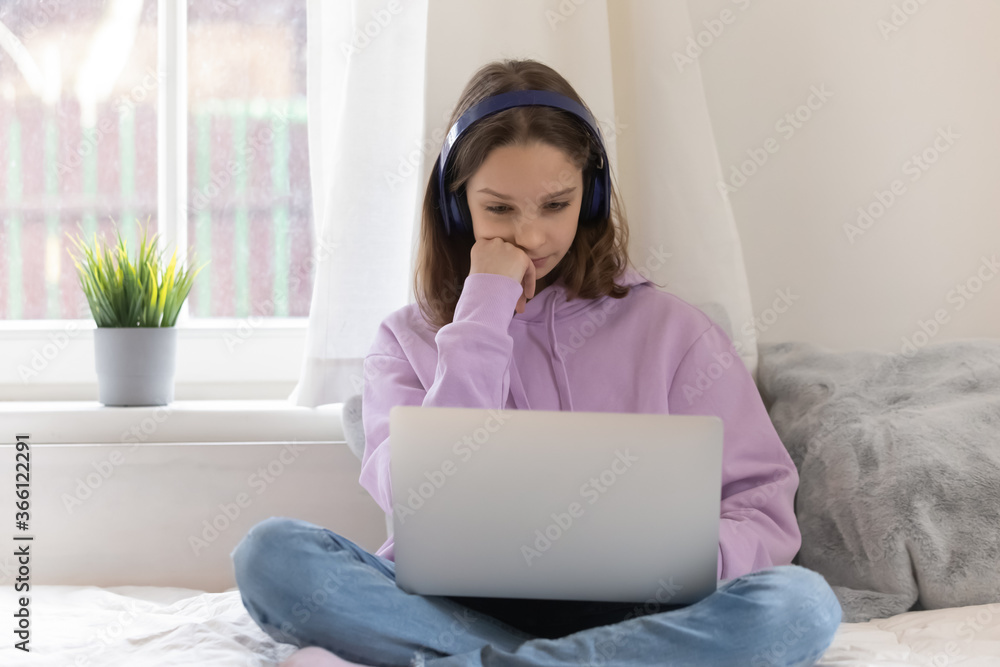 Bored teenage girl wearing headphones looking at laptop screen, sitting on bed at home, lazy schoolgirl studying online, watching boring webinar, educational course, distance learning concept