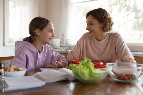 Happy mother and teenager daughter chatting, sitting at table in modern kitchen, smiling teen schoolgirl studying, working on school assignments, homework, mum cooking salad, preparing dinner © fizkes